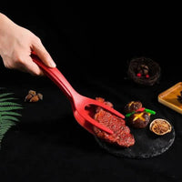 Person using a red multifunctional egg flipping shovel to handle cooked steak and vegetables on a black slate plate.