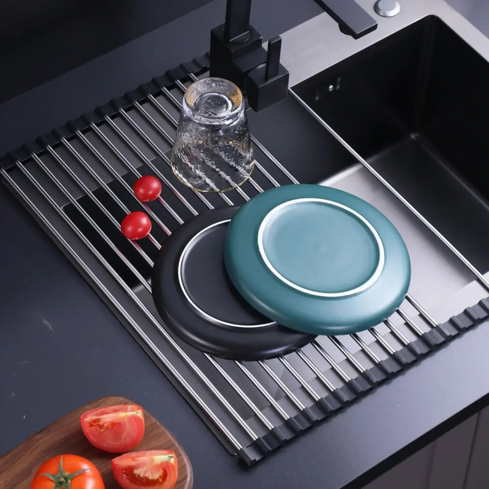 Foldable stainless steel draining rack in use with plates, glass, and tomatoes in a modern kitchen sink.