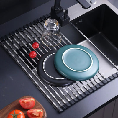 Foldable stainless steel draining rack in use with plates, glass, and tomatoes in a modern kitchen sink.